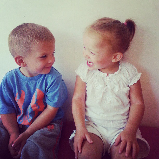 A young boy in a blue shirt and grey trousers sits next to a young girl in a white outfit with a ponytail. Both children are sitting against a plain light wall, smiling and looking happily at each other.