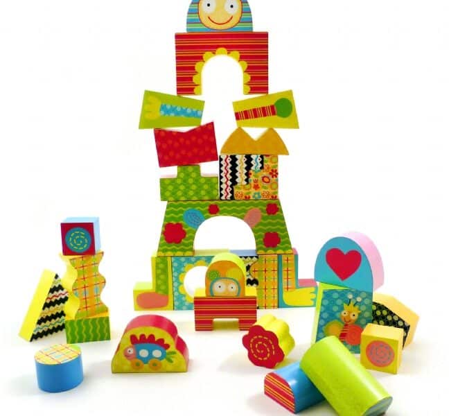 A colourful assortment of patterned wooden blocks is stacked in playful shapes, including rectangles, arches, circles, and triangles. Some blocks feature smiling faces, flowers, stripes, and hearts. Others are scattered around the base, inviting imaginative play.