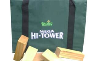 A green carrying bag labelled “Garden Games Mega Hi-Tower” sits upright. In front of it are five large, rectangular wooden blocks, some stacked and some lying flat, hinting at a giant outdoor Jenga-style game. The Garden Games logo is in the bottom right corner.