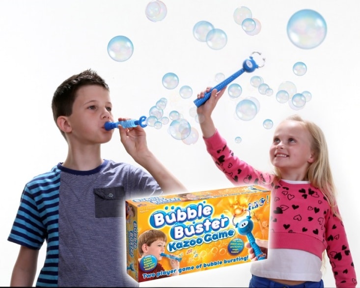 A boy and a girl stand against a white background, each blowing bubbles using blue bubble wands. Both look happy and engaged. There are bubbles floating around them. In the foreground is a box for the "Bubble Buster Kazoo Game.