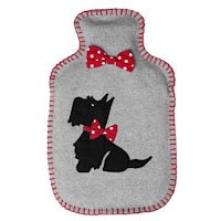A grey hot water bottle cover with red stitching, featuring an appliqué of a black dog adorned with two red, white-dotted bows—one below its neck and one above its head. The cover has rounded corners and a rectangular shape.