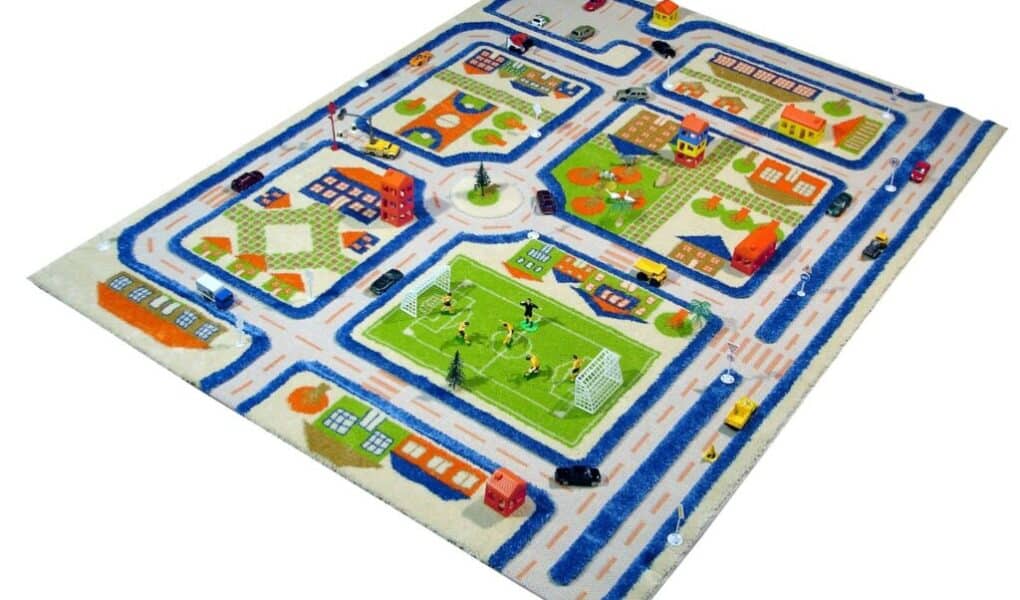 Children’s play rug showing a small town with winding blue roads, houses, buildings, cars, trees, a football pitch with players, and playgrounds. The rug has colourful residential and urban areas arranged in a neat, playful pattern.