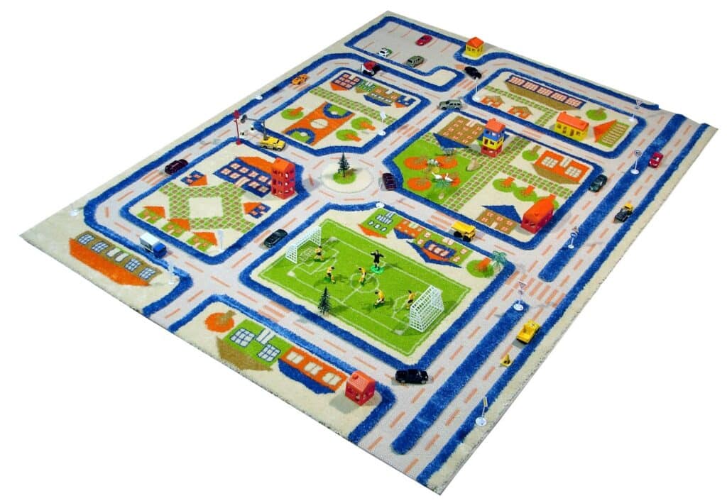 A colourful children’s playmat depicts a town with winding roads, houses, buildings, parks, a football pitch with players, trees, cars, and a playground, all illustrated in cartoon style on a rectangular rug.