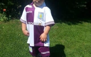 A young boy stands on green grass in bright sunlight, wearing a purple and white Aston Villa football kit with striped socks and purple sandals. His hands are on his hips and he looks confidently at the camera. Dense greenery is visible in the background.