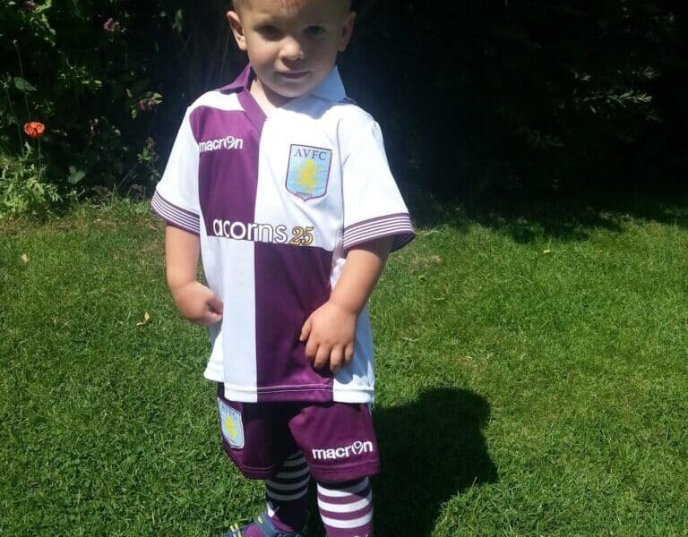 A young boy stands on green grass in bright sunlight, wearing a purple and white Aston Villa football kit with striped socks and purple sandals. His hands are on his hips and he looks confidently at the camera. Dense greenery is visible in the background.