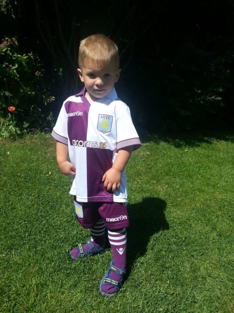 A young boy stands on green grass, wearing a purple and white Aston Villa football kit, including shirt, shorts, long socks, and blue sandals. He poses with one hand on his hip and faces the camera, with sunlight and shadows from nearby bushes behind him.