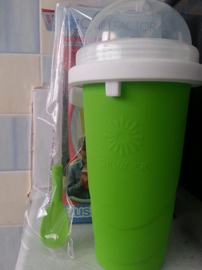 A green ChillFactor slush maker cup with a white rim and clear lid stands upright. A green spoon is sealed in plastic packaging beside it. Part of the product’s packaging, showing a child’s face, is visible in the background.