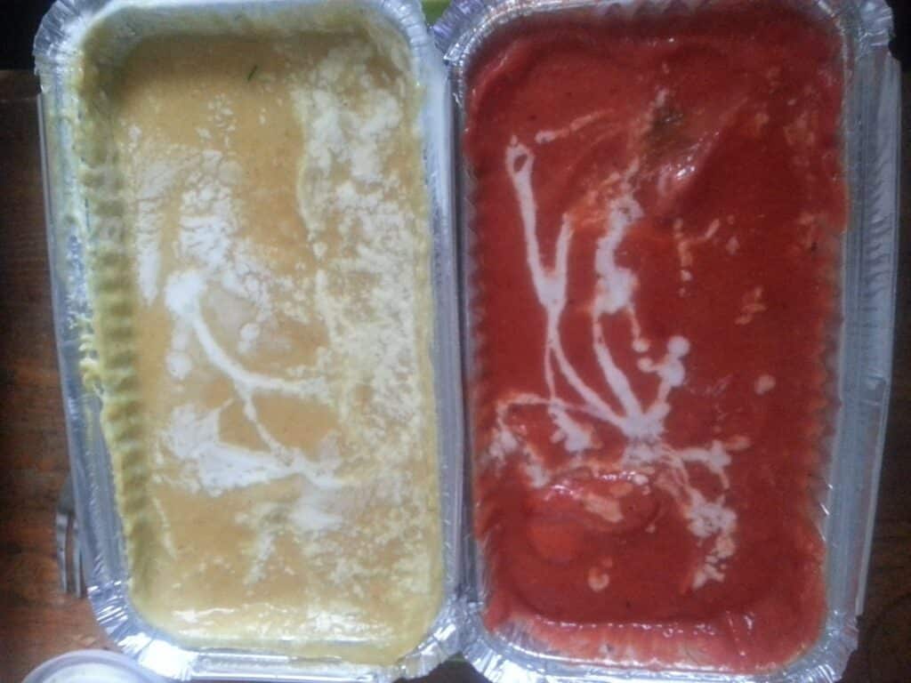 Two rectangular foil trays side by side: the left tray contains a creamy yellow curry with white streaks, and the right tray contains a rich red curry with similar white streak patterns. Both trays have smooth surfaces and appear freshly served.