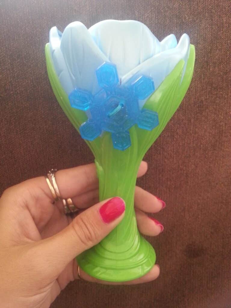A hand with red nail varnish holds a green plastic toy shaped like a flower, with pale blue petals and a blue snowflake decoration attached to the front. The background is a brown textured surface.
