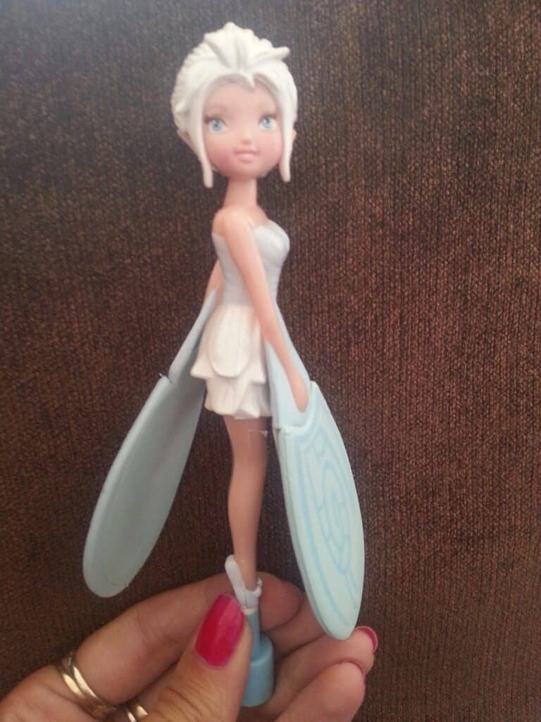 A hand with red-painted nails holds a doll with pale skin, white hair in an updo, and blue eyes. The doll wears a short, light blue dress and holds two large, light blue paddles in each hand. The background is a brown textured surface.
