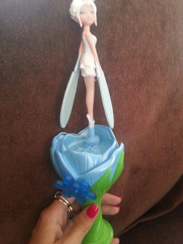 A hand with pink-painted nails holds a flying fairy toy with white hair, white dress, and pale blue wings, attached to a green base designed like a flower bud, against a brown fabric background.