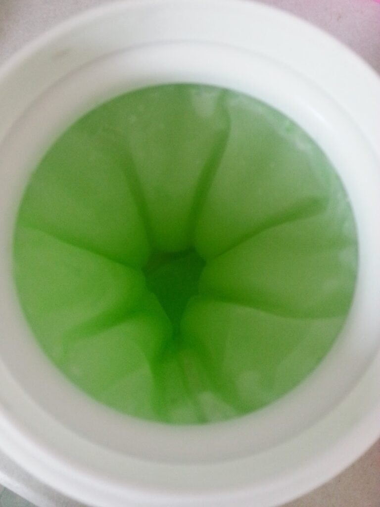 A close-up view of a white container filled with smooth, bright green gel, with the surface forming petal-like or wave-like shapes radiating from the centre. The gel appears glossy and semi-translucent.