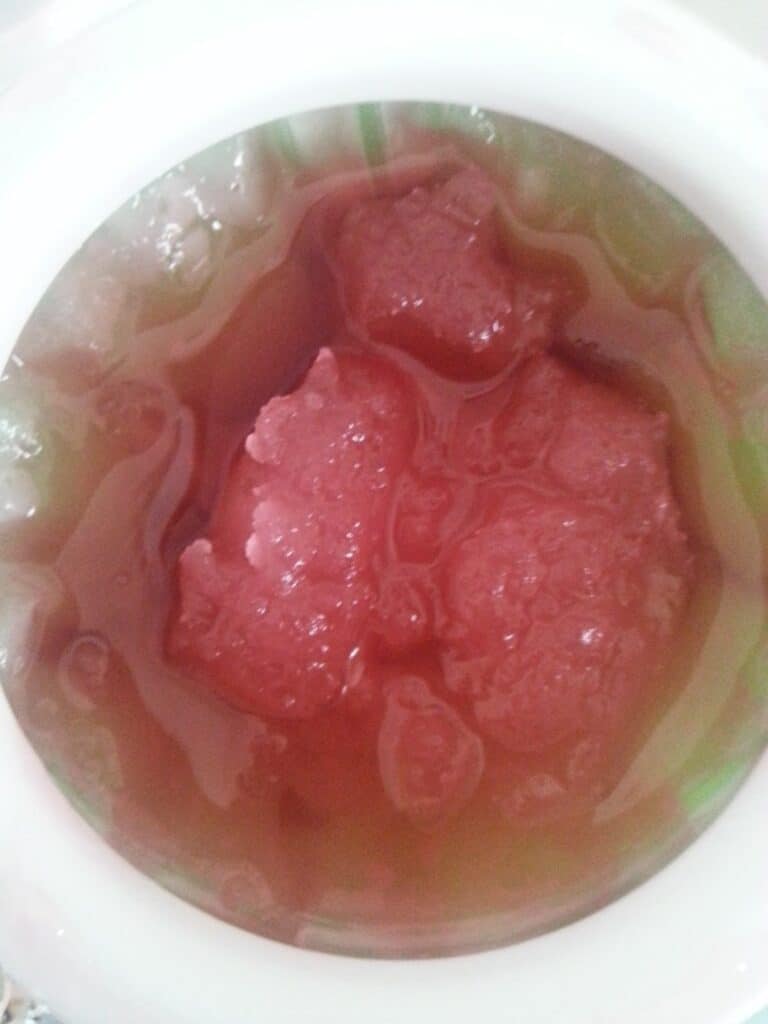 A close-up of a bowl containing reddish-pink ice chunks partially submerged in a translucent, light red liquid, possibly a dessert or drink, inside a white-rimmed container. The ice pieces have an irregular, slushy texture.