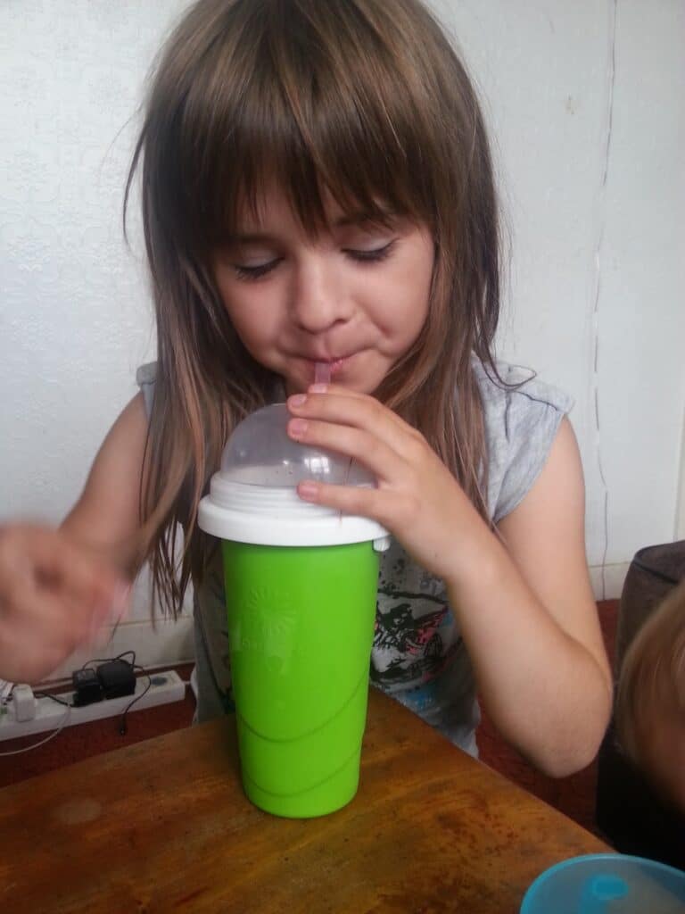 A young child with long brown hair sips from a pink straw in a large green cup with a clear dome lid, sitting at a wooden table. The child wears a sleeveless grey shirt. The background shows a white wall and an electrical socket with cables.