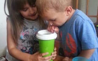 A young girl holds a green cup with a white lid while a young boy drinks from it using a straw. They are standing close together at a wooden table with a blue cup and a plastic lid nearby. The background includes a white wall and a wooden door with glass panes.