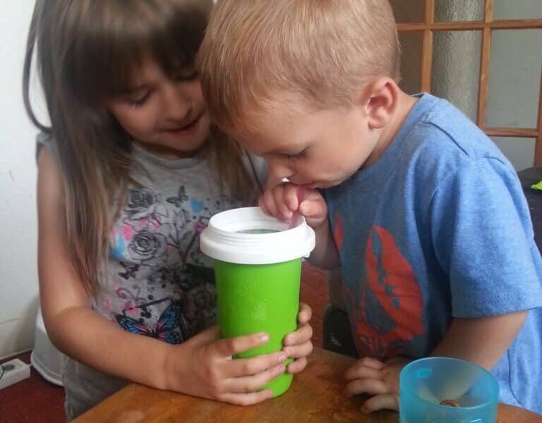 A young girl holds a green cup with a white lid while a young boy drinks from it using a straw. They are standing close together at a wooden table with a blue cup and a plastic lid nearby. The background includes a white wall and a wooden door with glass panes.