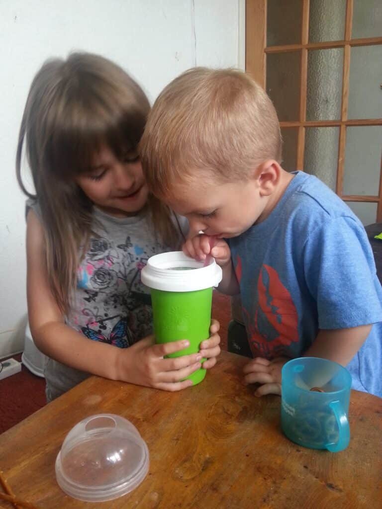 A young girl smiles while helping a younger boy drink from a green cup with a white lid. They both lean over the cup, which she holds with both hands. A blue cup and clear container lid sit on the wooden table beside them. They are indoors.