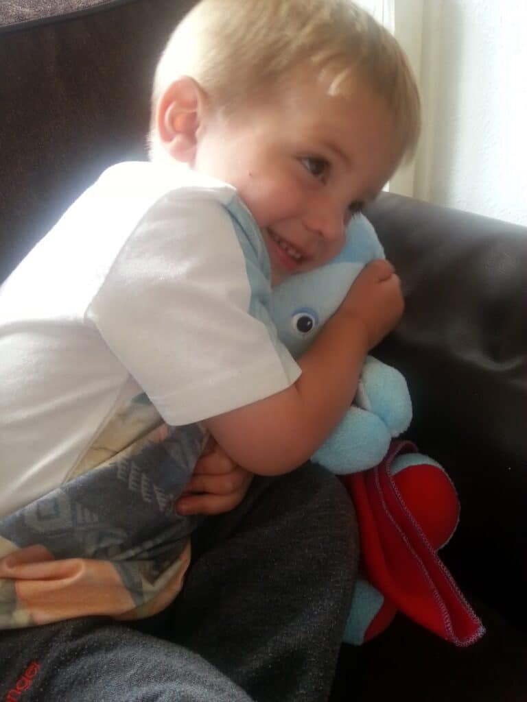A young child with blonde hair and a big smile cuddles a light blue soft toy while sitting on a dark sofa. The child looks happy and is wearing a white shirt and dark trousers, hugging the toy closely.