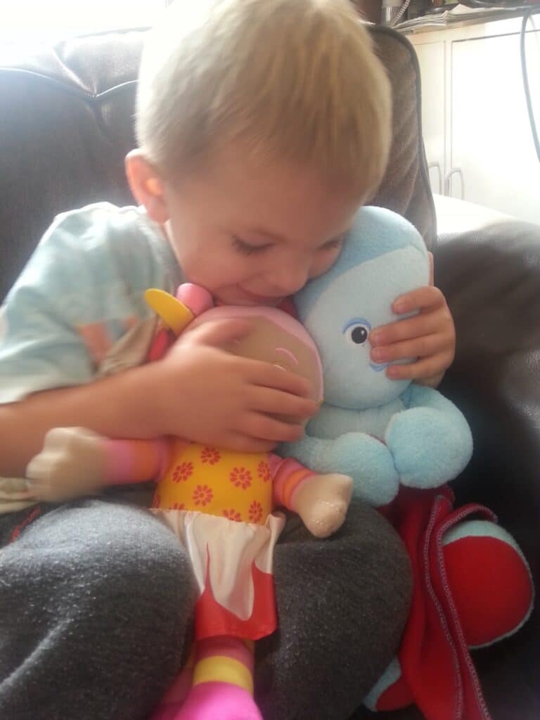A young child sits on a dark sofa, cuddling two soft toys—a pink and yellow doll and a light blue cuddly toy. The child smiles and hugs both toys close, appearing happy and content. Sunlight softly lights the scene.