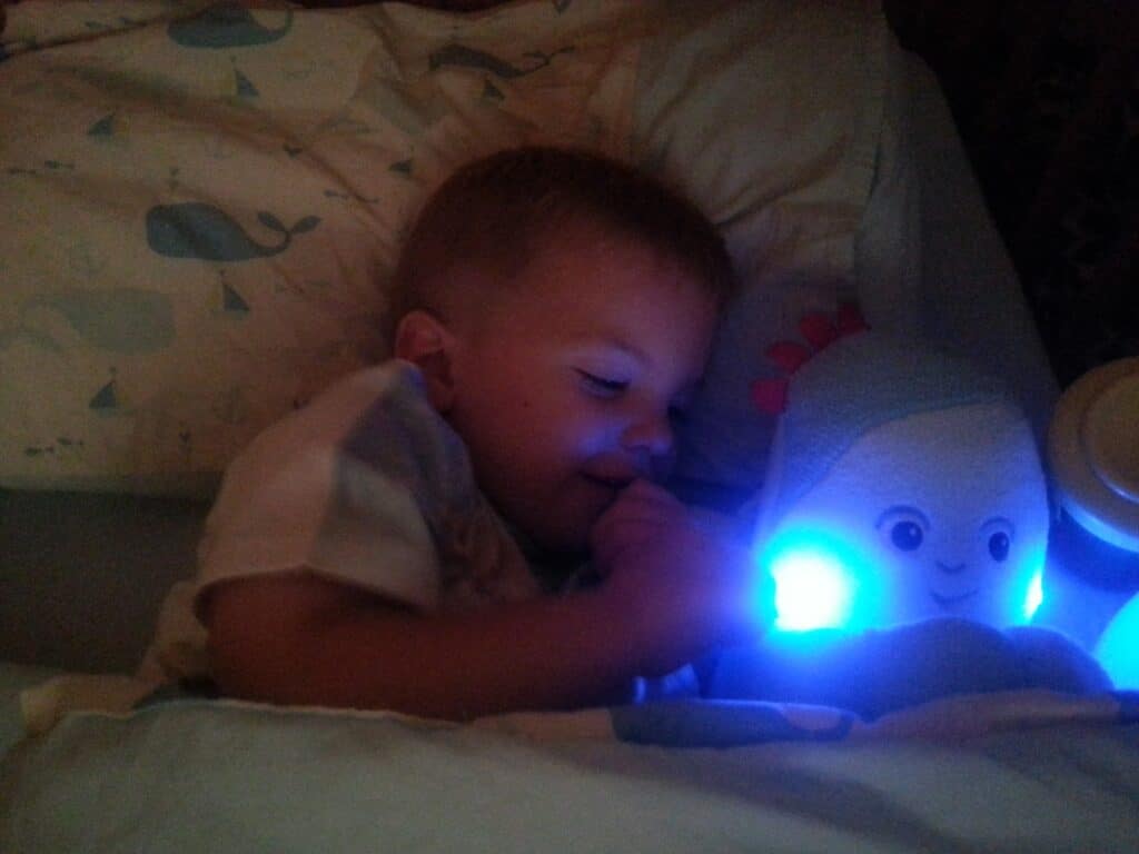 A young child lies in bed under a blanket with a blue whale pattern, holding a glowing blue soft toy that has a smiling face. The soft blue light from the toy illuminates the child's face as they cuddle it, ready to sleep.