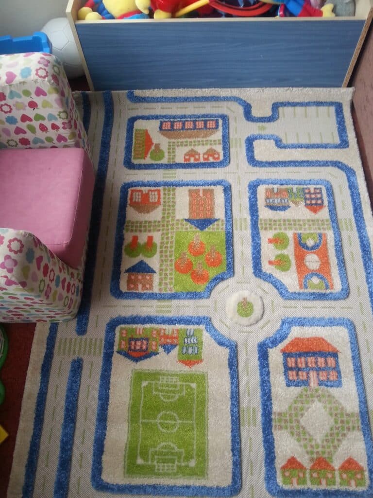 A colourful children's playmat features a cityscape with roads, houses, gardens, and a football pitch. A pink and floral-patterned armchair sits on the left, and a blue toy box filled with soft toys is visible at the top of the image.