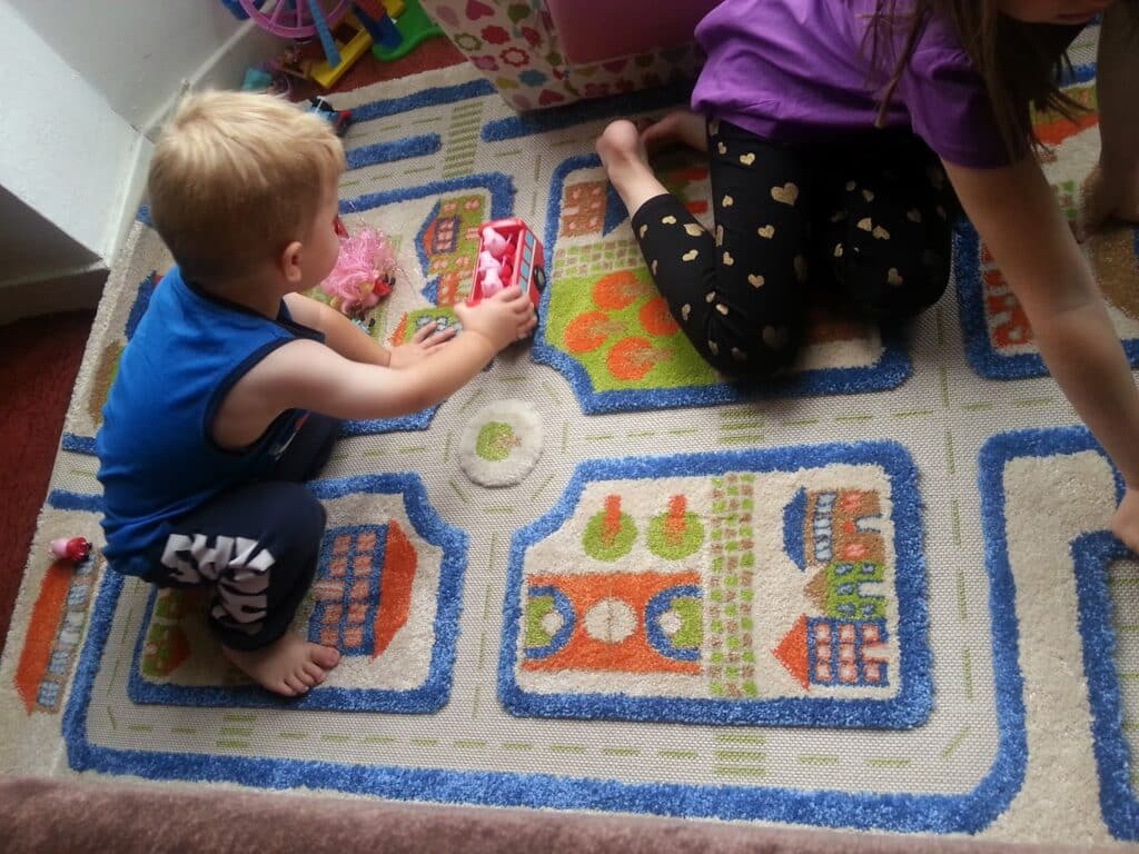 A young boy in a blue vest and a girl in a purple shirt with heart-patterned leggings play on a colourful rug with a town design. The boy pushes a toy fire engine, while the girl sits nearby with toys scattered around them.