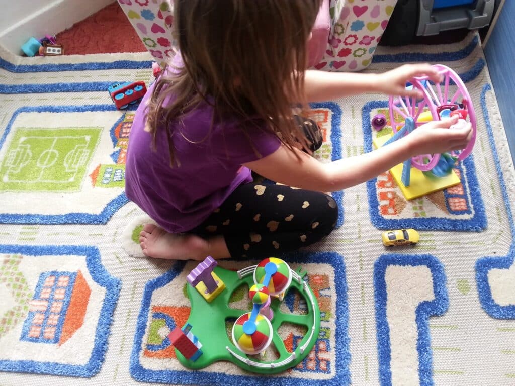 A young child with long brown hair, wearing a purple shirt and black leggings with gold hearts, sits barefoot on a colourful rug with a city and football pitch pattern, playing with a small pink Ferris wheel toy and other miniature plastic toys.