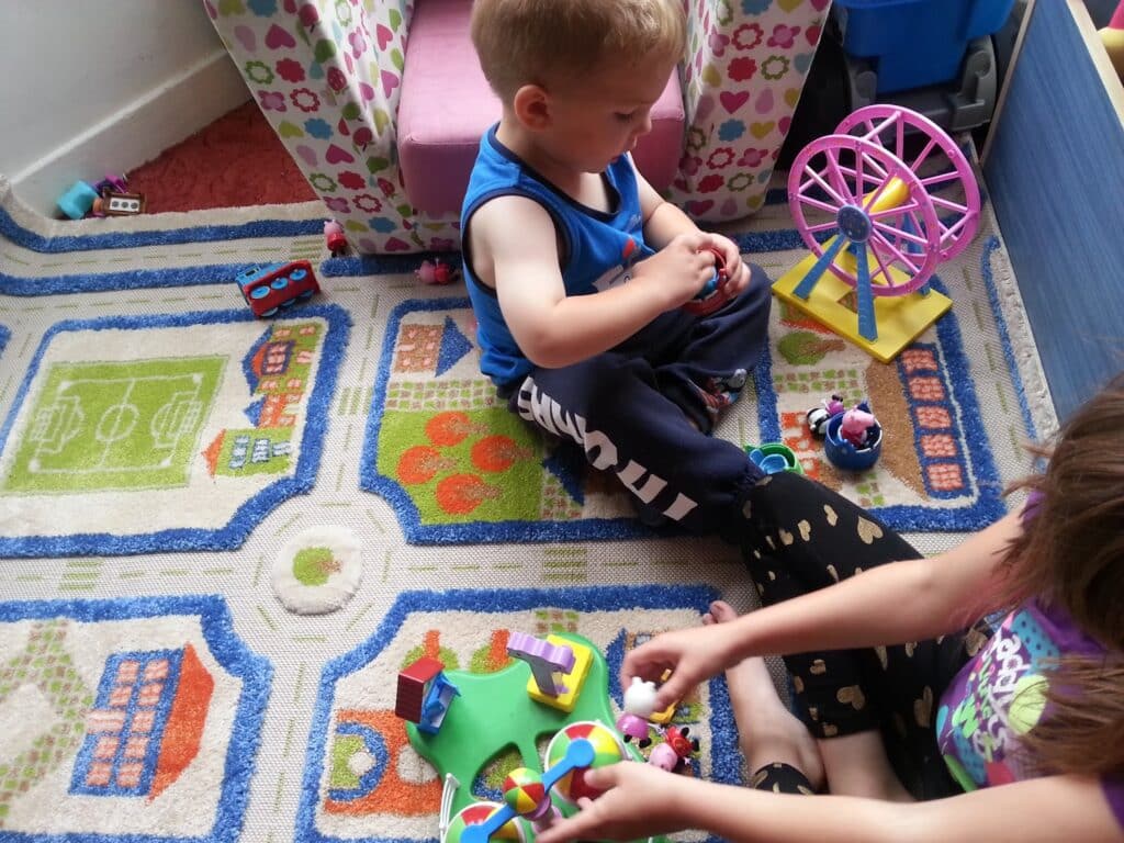 A young boy and girl sit on a colourful play rug decorated with sports pitches and roads. The boy is focused on small toys in his hands, while the girl plays with animal figurines and a toy Ferris wheel. A pink armchair is in the background.