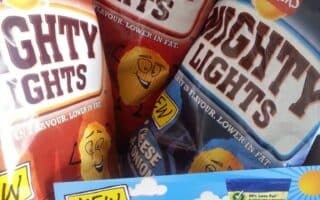 Three bags of Walkers Mighty Lights crisps—Roast Chicken, Cheese & Onion, and another flavour—are propped up behind a cardboard display showing all three flavours and advertising “30% less fat.” Cartoon crisp characters and bold “NEW” signs are visible.