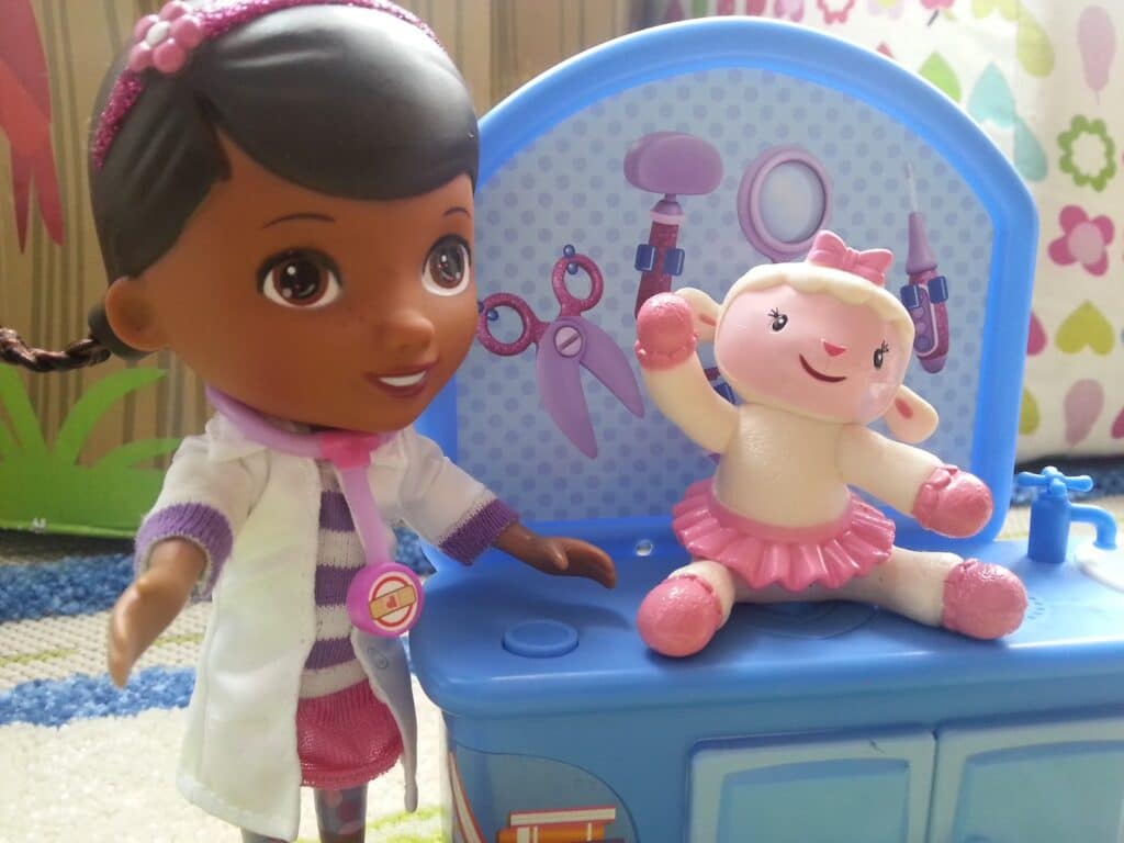 A dark-skinned doll dressed as a doctor with a pink hairband and stethoscope stands next to a smiling sheep toy in a pink tutu, sitting on a blue toy table with medical instruments illustrated on the background. Colourful designs decorate the surroundings.