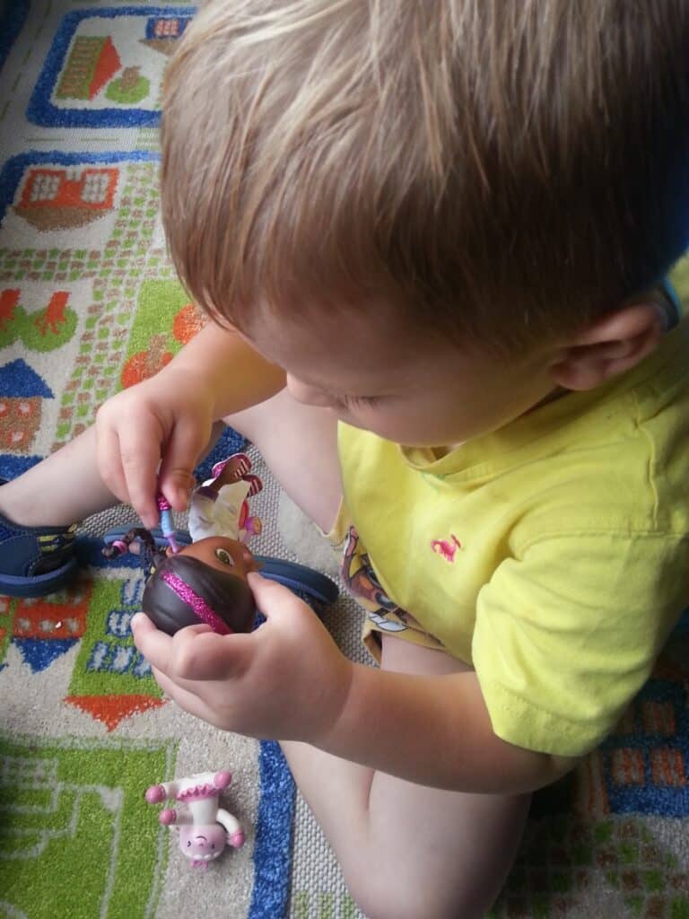 A young child with light brown hair wearing a yellow shirt sits on a colourful play rug, closely examining a small dark-skinned doll with a pink headband. Another small toy, resembling a lamb, lies nearby on the rug. The child appears focused.