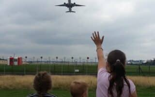 Three children stand outdoors, facing away from the camera, watching an aeroplane fly overhead. One child, in a pink shirt, raises their hand to wave at the plane. The sky is cloudy, and the scene is near an airport runway with grassy fields and airport equipment visible.