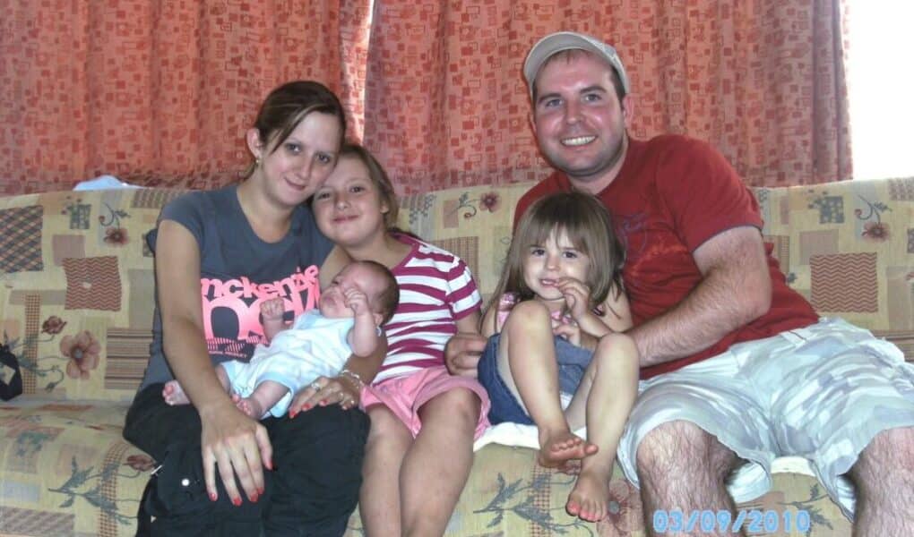 A family of five sits closely on a patterned sofa in front of orange curtains. The mother holds a newborn, with two young daughters beside her and a man in a red shirt and cap on the right, smiling. The date 03/09/2010 is on the bottom right.