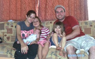 A family of five sits closely on a patterned sofa in front of orange curtains. The mother holds a newborn, with two young daughters beside her and a man in a red shirt and cap on the right, smiling. The date 03/09/2010 is on the bottom right.