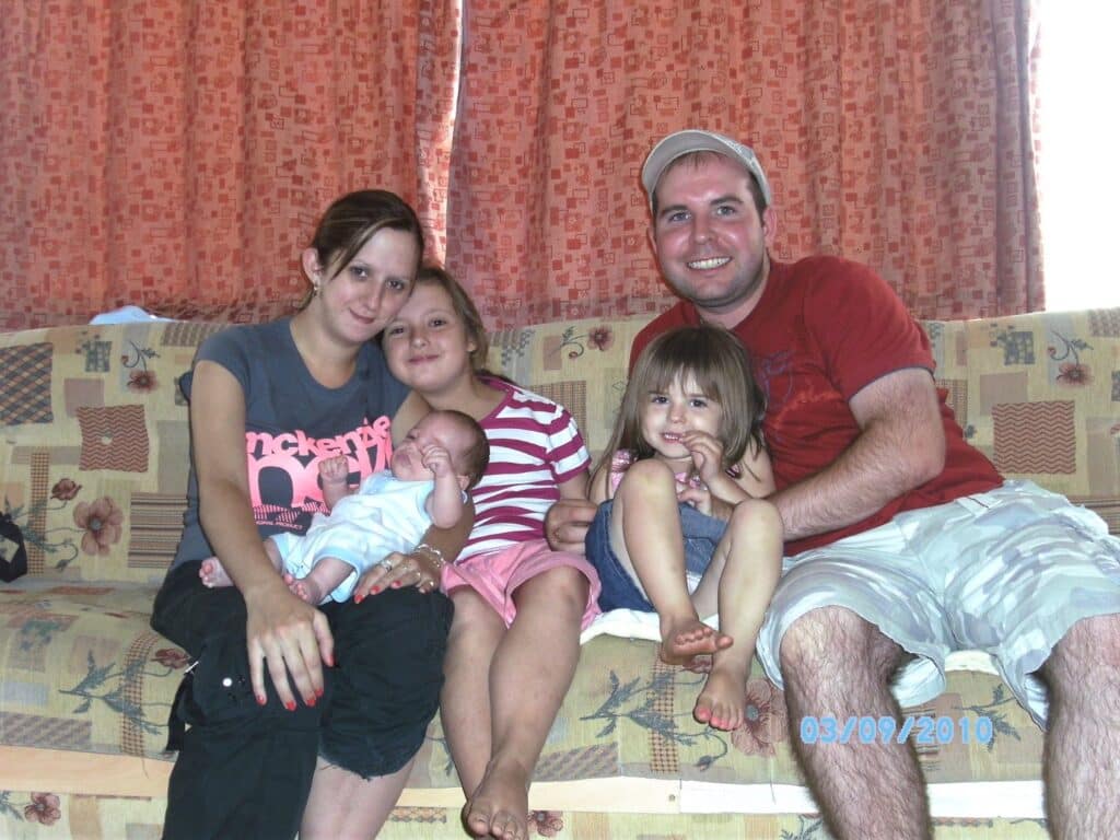A family of five sits on a patterned sofa. The mother holds a baby, with two young girls beside her. The father, wearing a cap and shorts, sits at the end, smiling. Red curtains are in the background. The date “03/09/2010” appears on the image.
