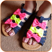 A pair of feet wearing dark blue strappy sandals with tan soles. Each sandal is decorated with three large bows—two bright pink and one neon green—attached to the straps. The person is standing on a brown carpet.