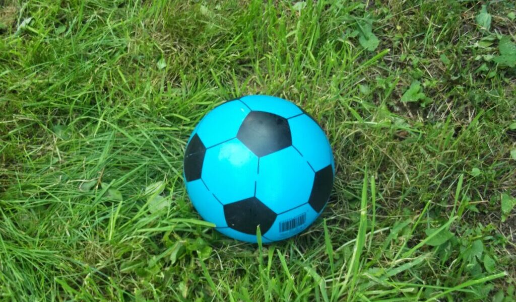 A bright blue and black football rests on uneven green grass. The grass varies in length, with some patches longer and some shorter. The ball’s barcode is visible on one of the blue panels near the bottom.