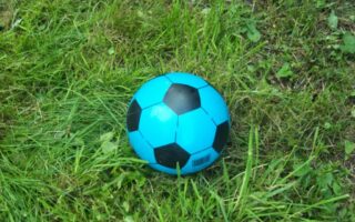 A bright blue and black football rests on uneven green grass. The grass varies in length, with some patches longer and some shorter. The ball’s barcode is visible on one of the blue panels near the bottom.