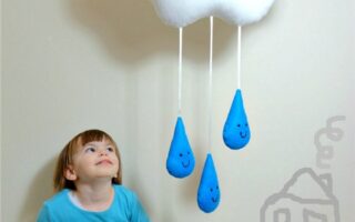 A young child in a blue shirt and patterned trousers sits on the floor, smiling up at a plush white cloud with a sleepy face. The cloud hangs on a wall with three blue raindrop soft toys dangling from it. A simple house drawing is on the wall beside the child.