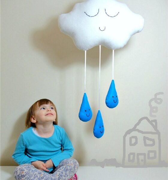 A young child in a blue shirt and patterned trousers sits on the floor, smiling up at a plush white cloud with a sleepy face. The cloud hangs on a wall with three blue raindrop soft toys dangling from it. A simple house drawing is on the wall beside the child.