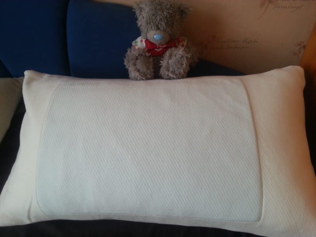 A large white pillow rests on a bed with a blue headboard. Behind the pillow, a grey plush teddy bear wearing a red bow sits upright. The wall in the background has faint writing or decorative script.