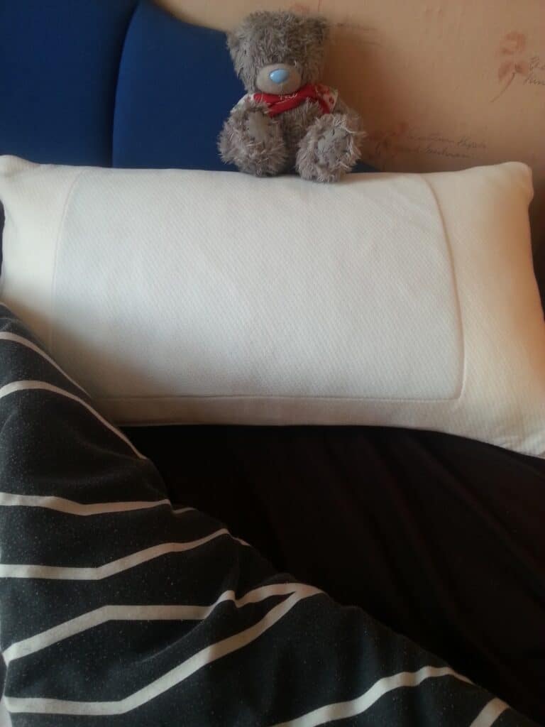 A grey teddy bear with a red scarf sits on a blue chair behind a large white rectangular cushion. In the foreground, there is a black and white striped blanket partially covering the lower part of the cushion.