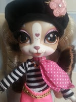 A doll with large brown and white eyes, a heart-shaped nose, and animal-like facial markings wears a black beret with a pink flower, a black-and-white striped top, a pink spotted scarf, and a pink skirt with a gold chain belt.