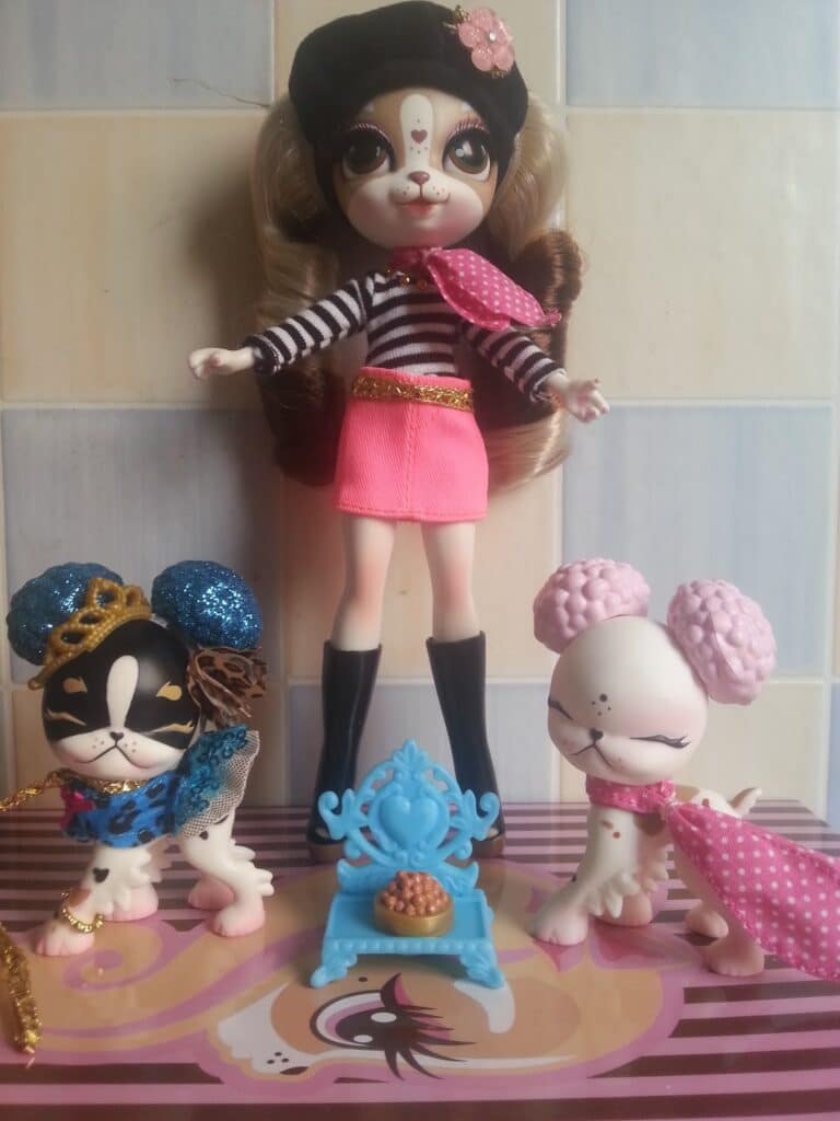 A doll with long hair, a black beret, striped top, pink skirt, and black boots stands in the centre. On either side are two small dog figurines, one black-and-white in blue accessories, the other white in pink. A blue chair with a pie sits between them.