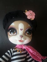 A toy dog with large brown eyes, white and brown fur, and a heart-shaped nose marking. It wears a black beret with a pink flower, a pink polka-dotted scarf, and a black-and-white striped shirt. The background is plain and dark.