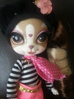 A doll with dog-like facial features, big brown eyes, and a heart-shaped nose wears a black beret with a pink flower, a black and white striped shirt, a pink scarf with white polka dots, gold necklace, and pink trousers with a gold belt.