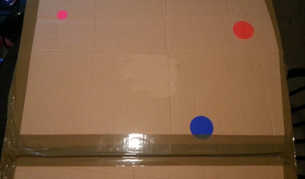 A large, taped cardboard box sits on a dark surface. The box has three round stickers on it: two red stickers at the top right and top left corners, and one blue sticker near the lower right side. The lighting is dim.