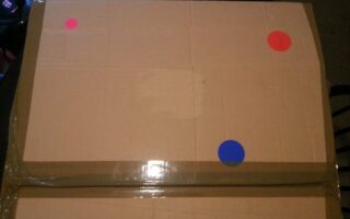 A large, taped cardboard box sits on a dark surface. The box has three round stickers on it: two red stickers at the top right and top left corners, and one blue sticker near the lower right side. The lighting is dim.