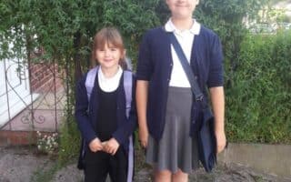 Two young girls stand outside in front of a hedge. Both wear navy cardigans, white shirts, and skirts; the younger girl has a dark dress, tights, and rucksack, while the older wears a grey skirt, white socks, and crossbody bag. They smile gently at the camera.