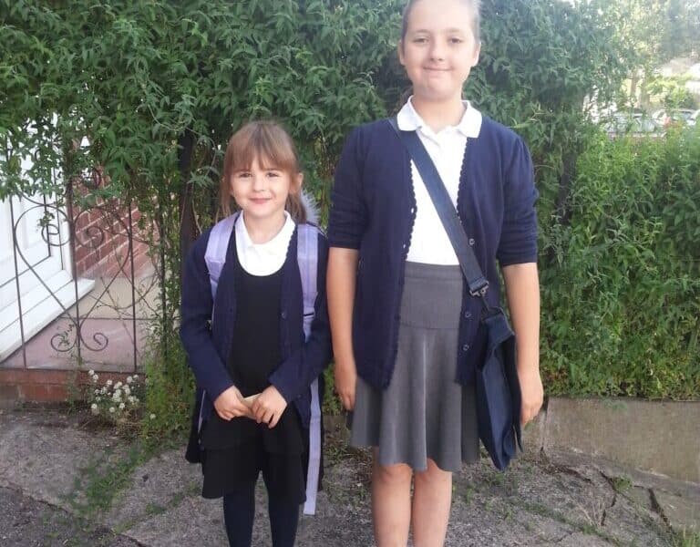 Two young girls stand outside in front of a hedge. Both wear navy cardigans, white shirts, and skirts; the younger girl has a dark dress, tights, and rucksack, while the older wears a grey skirt, white socks, and crossbody bag. They smile gently at the camera.